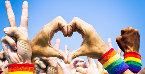 Hands forming a heart surrounded by other hands in the air making a peace sign and a justice fist. Their wrists have rainbow sweatbands.