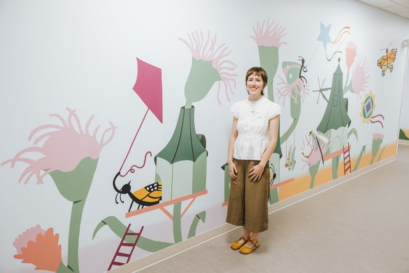 Grace LaPrade with her mural of small creatures and their houses among flowers