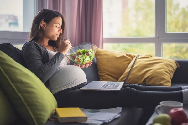 A pregnant woman eating a salad and using a computer